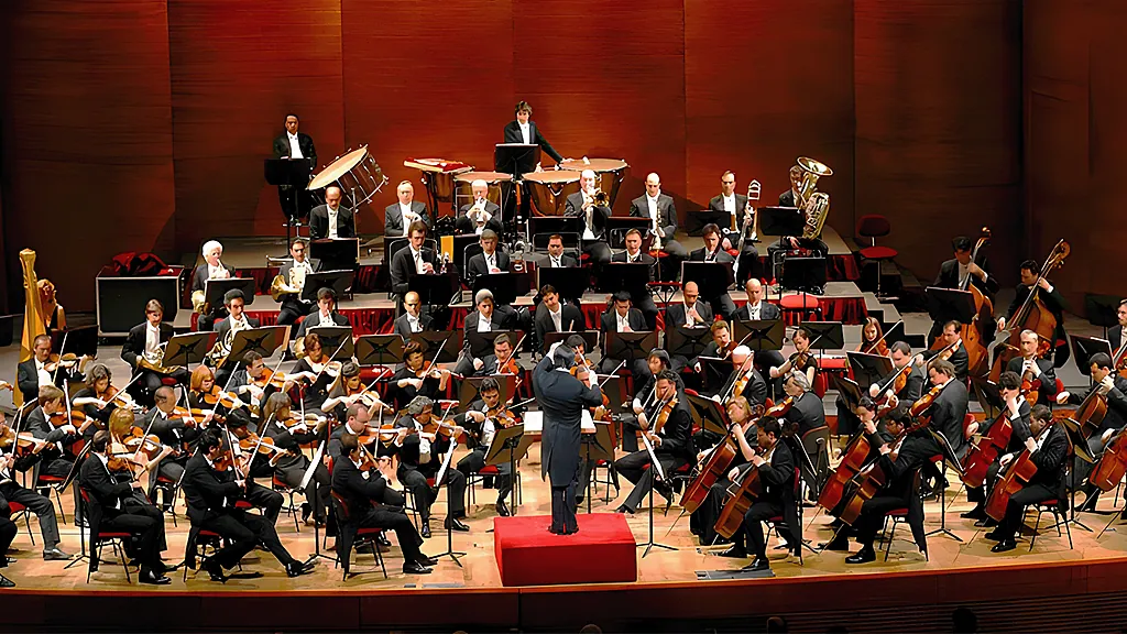 Una orquesta sinfónica completa en el escenario, con el director de pie sobre el podio frente a los músicos.